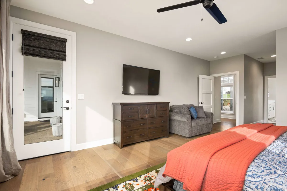 A comfortable bedroom is shown with a bright red blanket on the bed, a wooden dresser, a wall-mounted TV, a gray armchair, and a glass door leading to an outdoor area. The space feels airy and inviting, suitable for relaxation or enjoying morning routines.