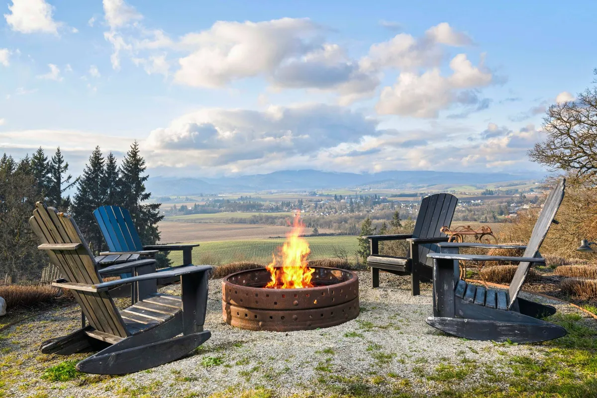 A cozy fire pit area surrounded by Adirondack chairs, set against the backdrop of rolling mountains and open sky. The golden hour lighting evokes warmth and invites gatherings for conversation, storytelling, or simply enjoying the sunset.