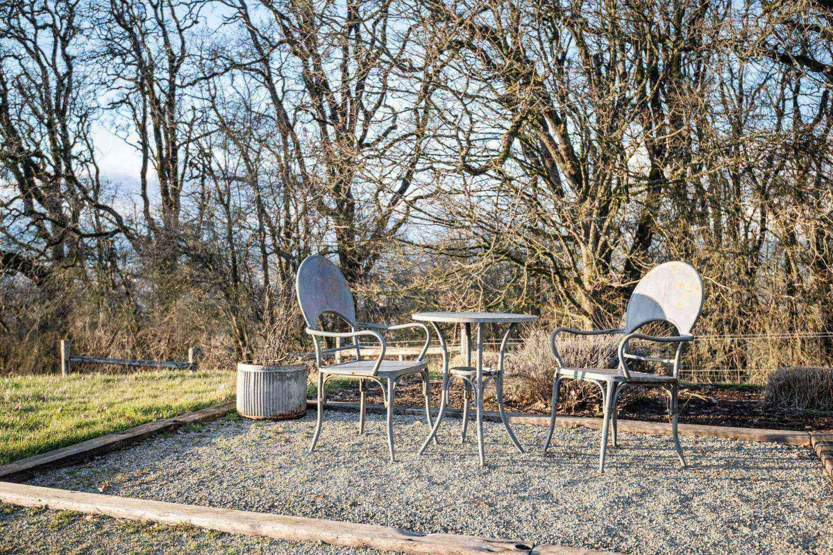 This peaceful outdoor setting includes two chairs and a small round table nestled among trees, creating a serene nook for quiet conversation or nature appreciation. The ambiance is intimate yet open to the surroundings, ideal for both group and individual retreat.