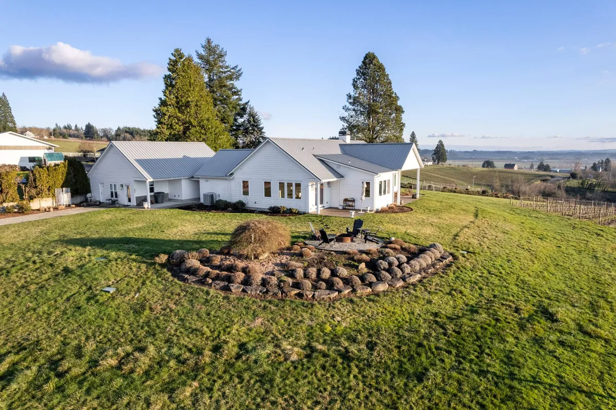 A landscaped backyard featuring a circular stone fire pit with cozy seating and lush green grass, framed by trees and the home in the background. The scene is tranquil, ideal for relaxed gatherings or quiet moments under the stars.