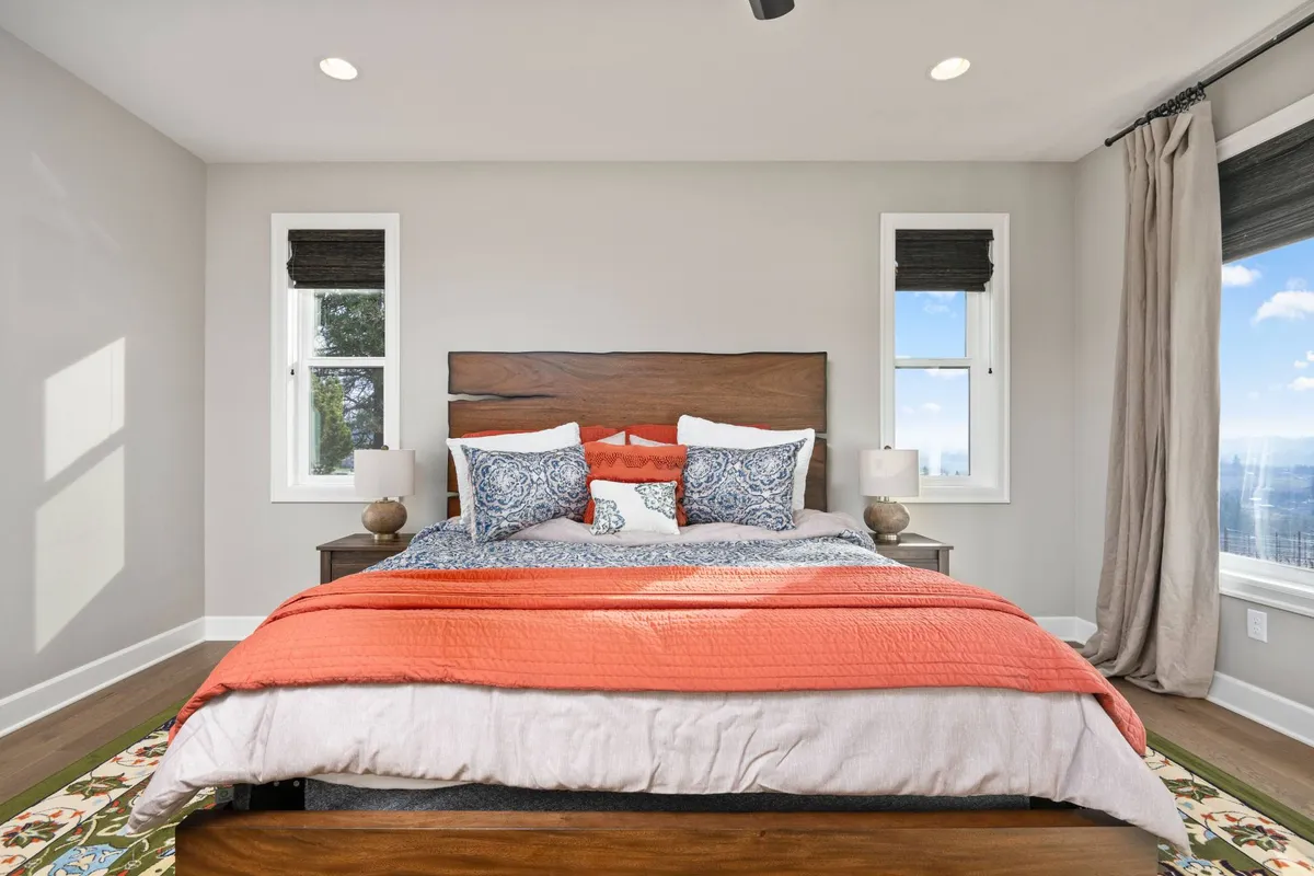 This image features a tranquil bedroom with a bed adorned in orange and blue bedding, flanked by decorative lamps on nightstands and large windows that welcome abundant natural light. The setting suggests a peaceful retreat designed for relaxation or enjoying quiet moments, such as reading or savoring a morning coffee.