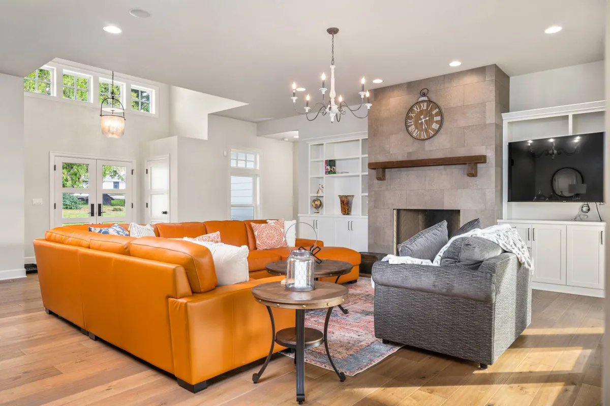 A welcoming living room centered around an orange sectional sofa, a fireplace, and a stylish chandelier. Decorative shelves and a TV unit add to the sense of thoughtful design, while the warm lighting and open plan invite relaxation and socializing in a calm setting.