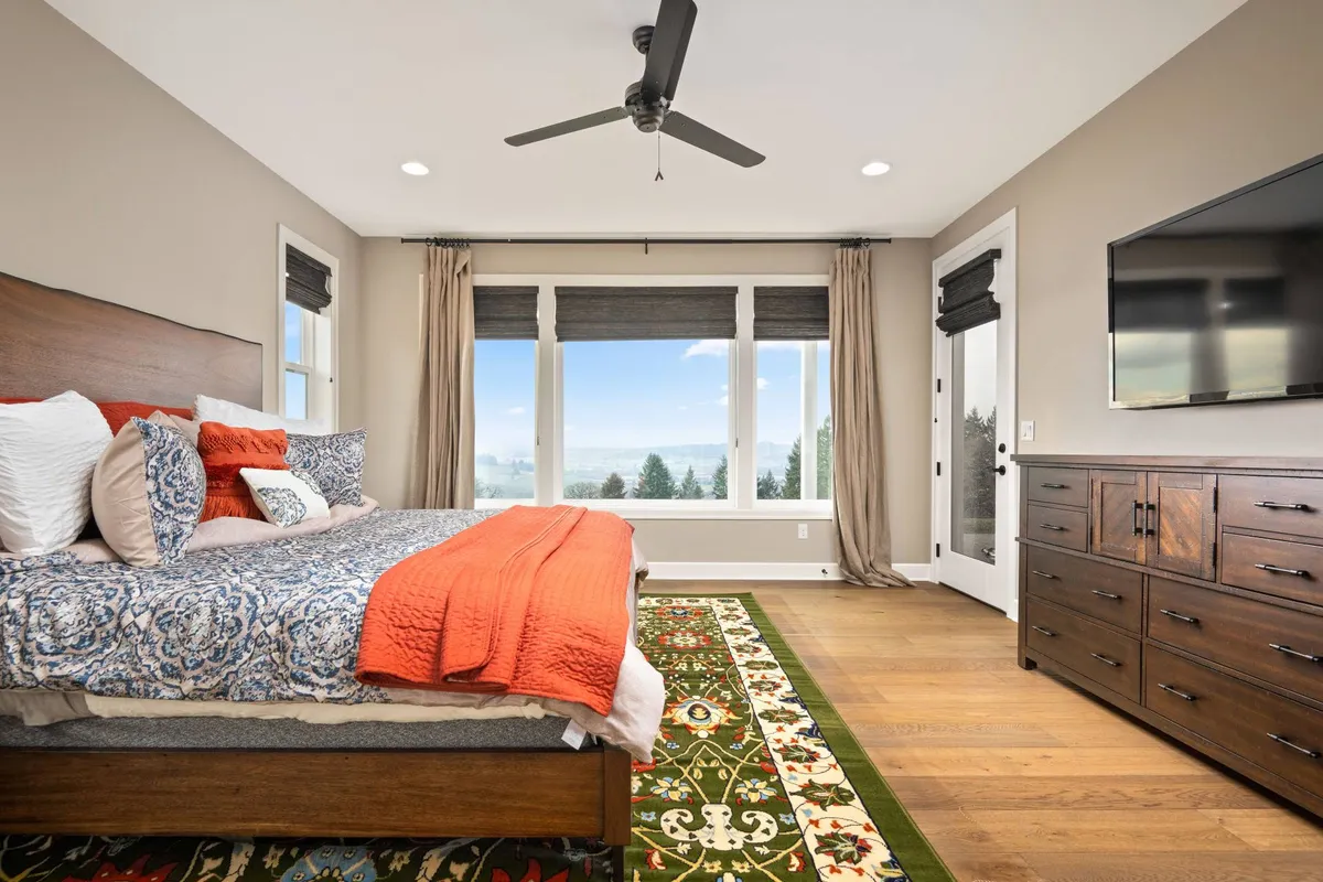 A spacious bedroom featuring a bed with colorful bedding, a decorative rug, and a wooden dresser. Large windows provide ample natural light and offer beautiful views, while a wall-mounted TV adds an element of comfort and leisure. The room exudes a cozy, peaceful ambiance ideal for relaxation and quiet reflection.