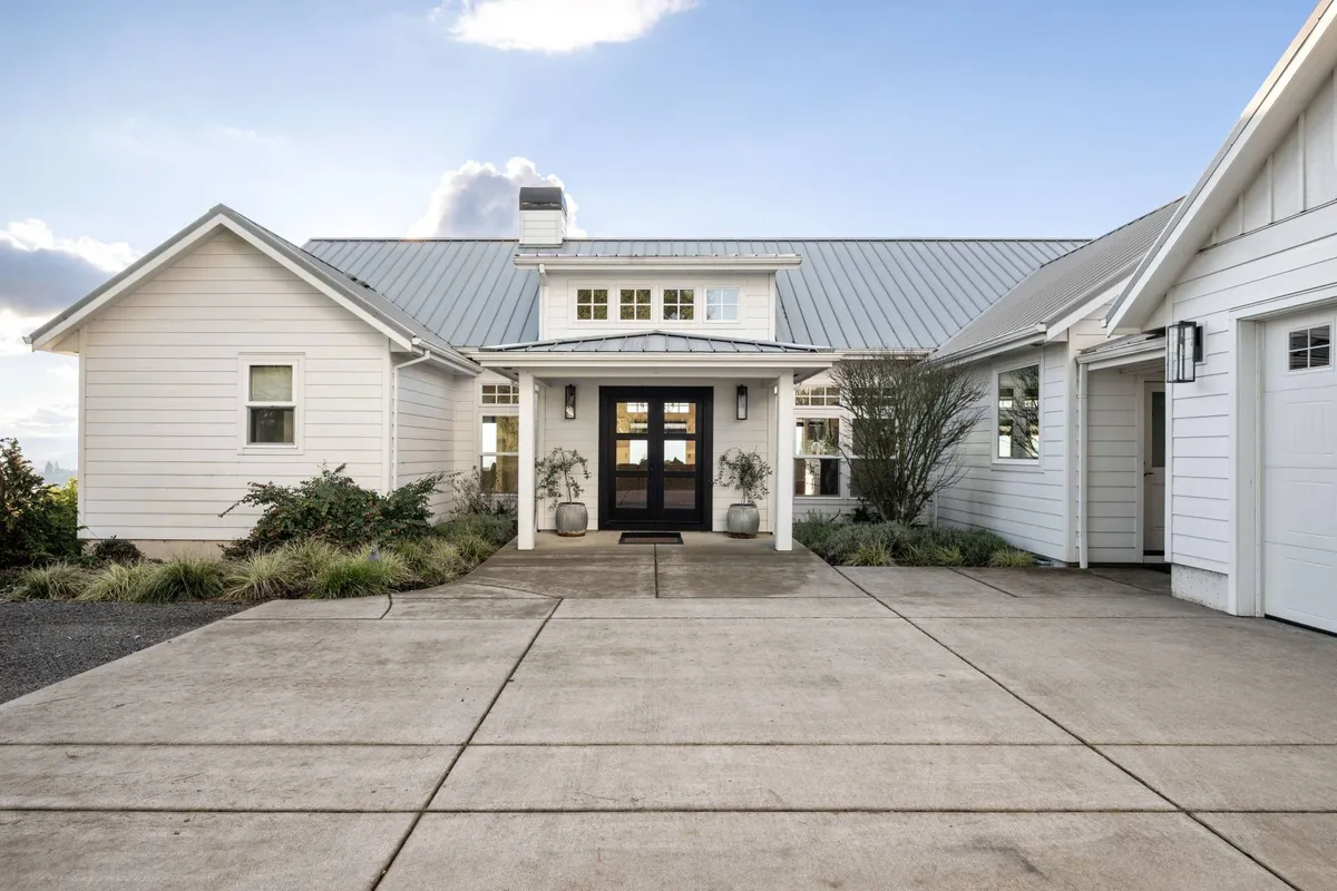 The image features the estate’s inviting front entrance framed by beautiful landscaping and a welcoming driveway. The house stands as a gateway to both comfort and the region’s renowned wineries, representing the hospitality and curb appeal of the property.