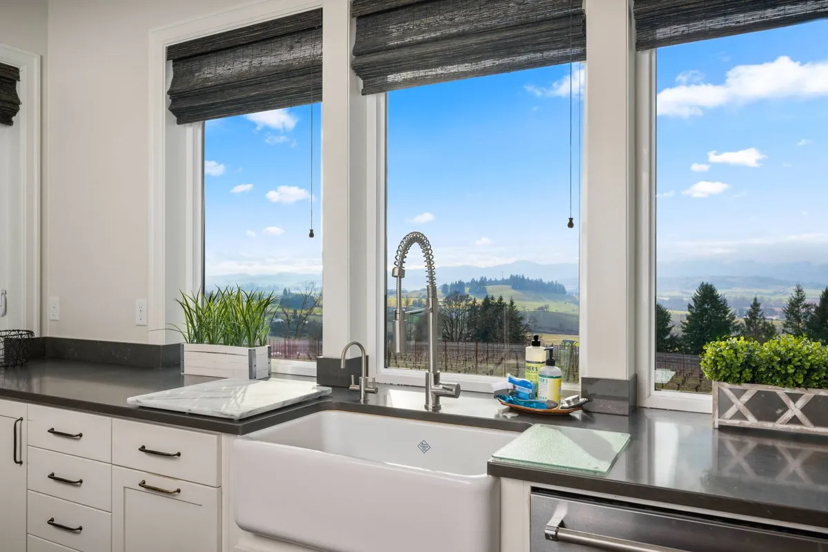 A sunlit kitchen space with expansive windows framing vineyard or countryside views. Sleek modern fixtures and a countertop decorated with subtle accessories create a refreshing, open atmosphere ideal for slow mornings or serene evenings.