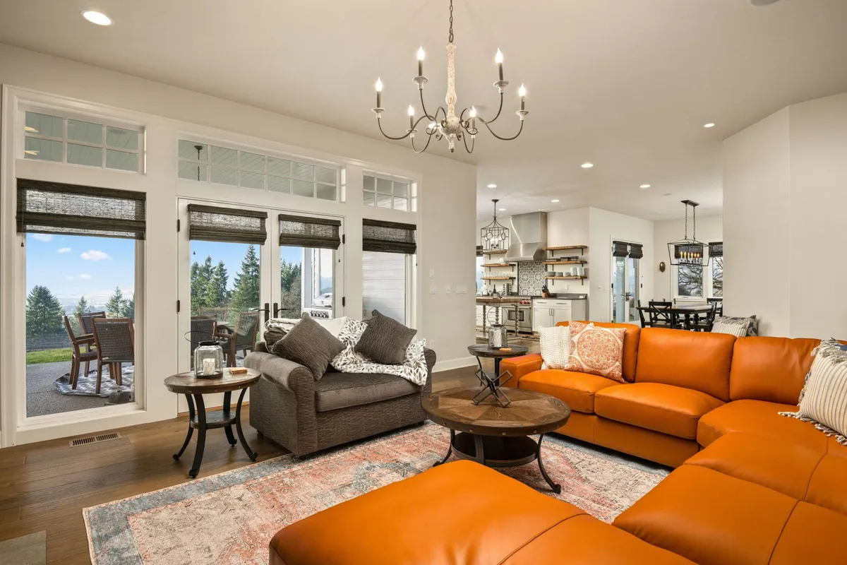 The cozy living area is centered around a bold orange sectional sofa and a round coffee table, set beneath a sparkling chandelier. Large windows bathe the space in natural light and offer breathtaking views of the surrounding landscape, reinforcing a sense of wine country serenity. The arrangement encourages socializing and relaxation.